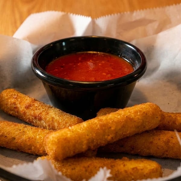 Golden-brown mozzarella sticks and a small black bowl of red marinara sauce in a basket.