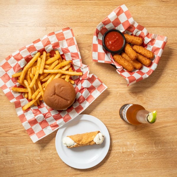 Overhead view of a burger, fries, mozzarella sticks, a cannoli, and a beer on a wooden table.