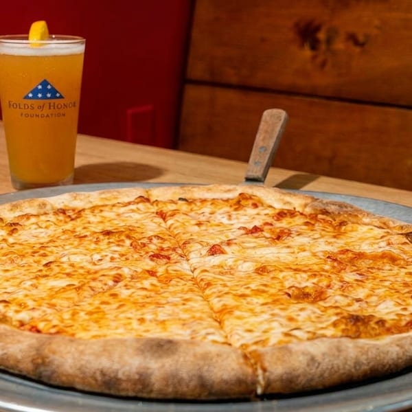 A whole cheese pizza on a metal pan next to a glass of beer with a lemon wedge.