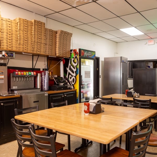Interior of a casual restaurant with wooden tables, black chairs, soda fountain, and stacked pizza boxes.