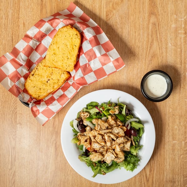 Overhead view of a chicken salad with dressing and two slices of garlic bread on a wooden table.