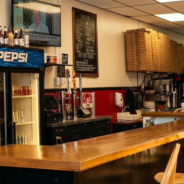 Interior of a casual restaurant with a wooden bar, bar stools, Pepsi fridge, and pizza boxes.