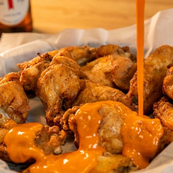 Crispy fried chicken wings in a basket, with orange sauce being poured over them, and a beer bottle in the background.