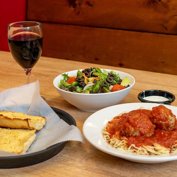 Spaghetti and meatballs, a side salad, garlic bread, and a glass of red wine on a wooden table.