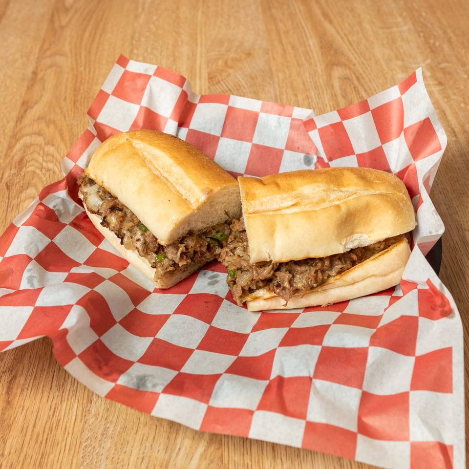 Cheesesteak sandwich cut in half, filled with meat, onions, and peppers on checkered paper.