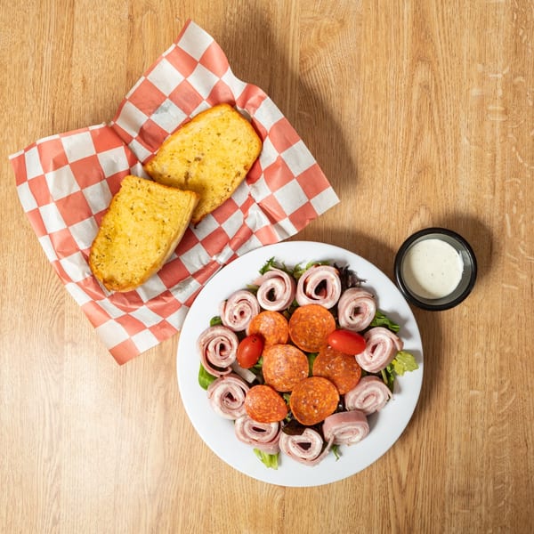 Antipasto salad with pepperoni, ham rolls, tomatoes, and garlic bread on a wooden table.