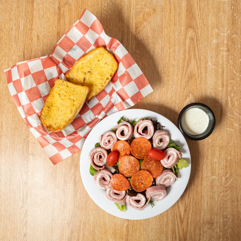 Antipasto salad with pepperoni, ham rolls, tomatoes, and garlic bread on a wooden table.