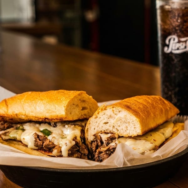 Cheesesteak sandwich cut in half, with melted cheese and peppers, next to a glass of Pepsi.